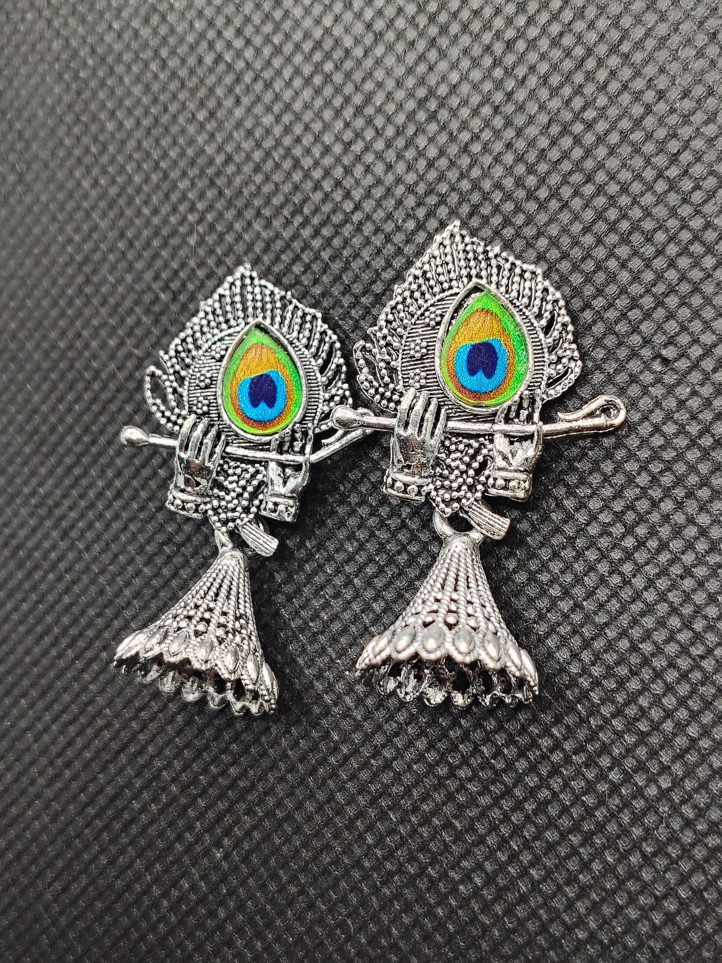 Pair of silver fish-shaped earrings with peacock feather details on a textured gray background